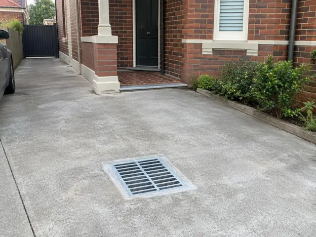Clean outdoor drain grate set in a concrete driveway beside a brick house in Dulwich Hill